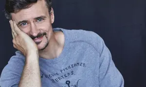 Mateusz Dela zamyślony, opiera głowę na dłoni. Na szarym swetrze widnieje napis "WELCOME FISHERMEN, HUNTERS".