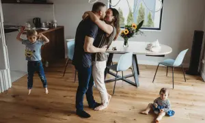 Rodzice w uścisku, starszy syn skacze, młodsza córka siedzi na podłodze. Urocza sesja lifestyle w domu.