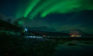 Nocne niebo rozświetla zorza polarna. Na pierwszym planie widać fotografa z lampą, który robi zdjecie z fleszem.