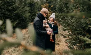 Rodzina podczas sesji świątecznej w plenerze. Ojciec trzyma dziecko, matka uśmiecha się, otoczeni choinkami.