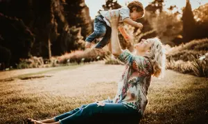 Mama rzuca dzieckiem w górę podczas śmiesznej sesji rodzinnej w parku. Słońce zachodzi, tworząc ciepłą, złotą poświatę.