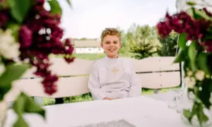 Uśmiechnięty chłopiec w albie, gotowy na sesję komunijną. Siedzi na ławce w otoczeniu kwitnących bzów.