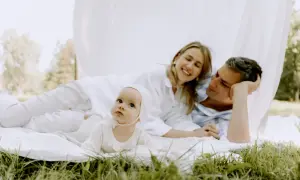 Urocza sesja z niemowlakiem w plenerze. Rodzice leżą na kocu, a dziecko w białym ubranku patrzy w obiektyw.