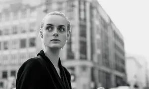 Monochromatyczna fotografia. Młoda kobieta o blond włosach patrzy w bok, na tle miejskiej architektury.