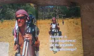 Piotr Trybalski z aparatem i statywem w gorącej Afryce. Obok niego kobieta i dziecko na pustynnej drodze.