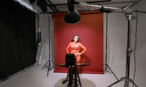 Modelka w pomarańczowej sukience pozuje podczas sesji studyjnej. W tle czerwone tło, oświetlenie i sprzęt fotograficzny.