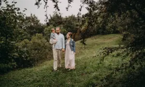 Rodzina podczas sesji rodzinnej w plenerze latem. Ojciec trzyma córeczkę, matka idzie obok.
