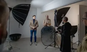W studiu fotograficznym trwa sesja. Widać ludzi, sprzęt i modyfikatory światła w fotografii, w tym parasole i blendę.