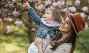Uśmiechnięta mama w kapeluszu trzyma dziecko podczas sesji wiosennej dzieci. Maluch wskazuje palcem na kwitnącą magnolię.