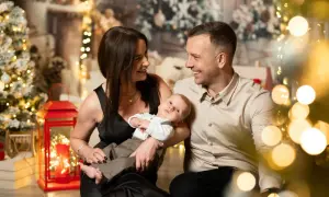 Rodzina podczas sesji bożonarodzeniowych. Mama i tata patrzą na siebie z uśmiechem, trzymając niemowlę. W tle choinki i lampki.
