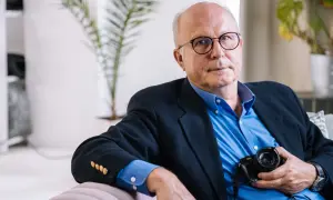 Tomasz Tomaszewski, w okularach i granatowej marynarce, trzyma aparat fotograficzny.