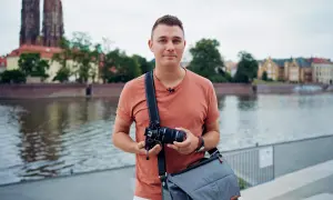 Mężczyzna z aparatem i torbą na ramieniu, gotowy do robienia zdjęć na dworze. W tle rzeka i zabytkowe budynki.