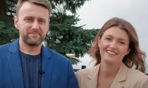 Uśmiechnięty adam kozioł w niebieskiej marynarce obok kobiety w beżowej.