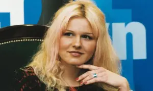 Agnieszka Lorek z blond włosami i delikatnym uśmiechem, oparta o fotel, z ręką podtrzymującą podbródek.