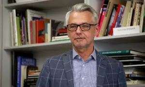 Andrzej Grygiel w okularach, w kratowaną marynarkę, na tle regałów z książkami o sztuce i fotografii.