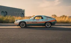 Klasyczna fotografia samochodowa: Porsche 928 w kolorach retro, z numerem 28 i logiem Brightlog, na tle nowoczesnego budynku i złotej trawy.