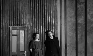 Dwoje aktorów w czerni, w dramatycznej pozie, na tle drewnianej ściany i drzwi. Ta fotografia teatralna uchwyciła moment napięcia.