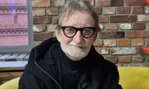 Maciej Mańkowski w okularach, z siwą brodą, w czarnej bluzie na tle ceglanej ściany.