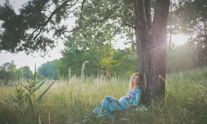 Piękna sesja ciążowa w letnim krajobrazie. Kobieta w kwiecistej sukience odpoczywa pod drzewem, otoczona wysoką trawą.