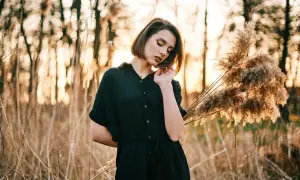 Młoda kobieta w czarnej sukience pozuje na tle zachodzącego słońca i trzcin. Idealna inspiracja na kurs fotografii portretowej.