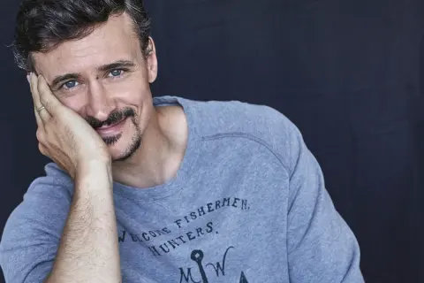 Mateusz Dela zamyślony, opiera głowę na dłoni. Na szarym swetrze widnieje napis "WELCOME FISHERMEN, HUNTERS".