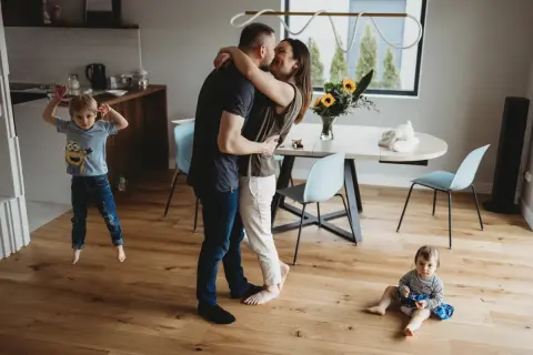 Rodzice w uścisku, starszy syn skacze, młodsza córka siedzi na podłodze. Urocza sesja lifestyle w domu.
