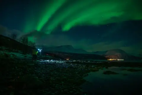 Nocne niebo rozświetla zorza polarna. Na pierwszym planie widać fotografa z lampą, który robi zdjecie z fleszem.