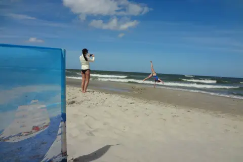 Dziewczyna robi zdjęcie akrobatce na plaży. W tle widać morze i chmury, a horyzont jest lekko krzywy.