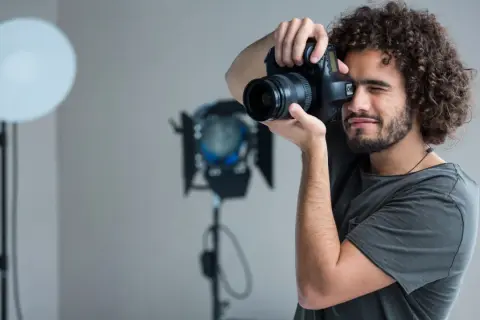 Mężczyzna z kręconymi włosami i aparatem w dłoniach, ćwiczący, jak zostać fotografem. W tle lampy studyjne.