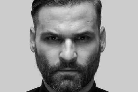 Intensywne spojrzenie Karola Grygoruka w czarno-białym kadrze. Mężczyzna z zarostem i zaczesanymi włosami patrzy prosto w obiektyw.
