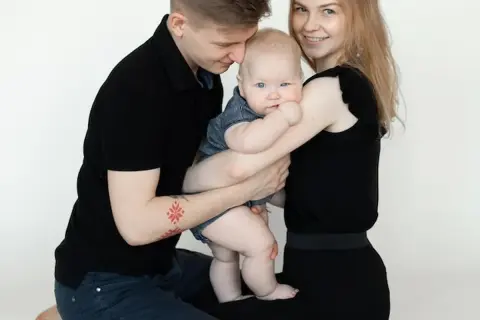Szczęśliwa rodzina podczas sesji z niemowlakiem. Tata tuli dziecko, mama uśmiecha się.