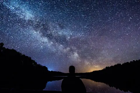 Sylwetka człowieka patrzy na Drogę Mleczną nad jeziorem. Idealny kadr, by pokazać, jak fotografować gwiazdy.