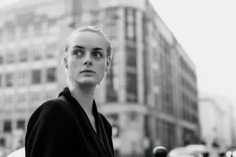Monochromatyczna fotografia. Młoda kobieta o blond włosach patrzy w bok, na tle miejskiej architektury.