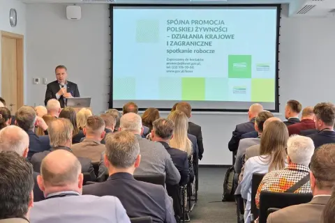 Mężczyzna przemawia na spotkaniu roboczym. Na ekranie widnieje napis "Spójna promocja polskiej żywności". Może to być wada obiektywu, która lekko zniekształca obraz.