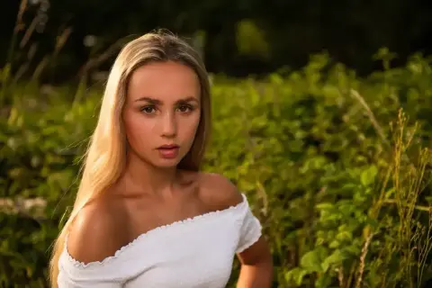 Młoda kobieta z długimi blond włosami, ubrana w białą, odkrywającą ramiona bluzkę, patrzy prosto w obiektyw na tle zielonej, rozmytej roślinności.