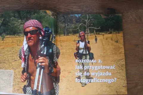Piotr Trybalski z aparatem i statywem w gorącej Afryce. Obok niego kobieta i dziecko na pustynnej drodze.