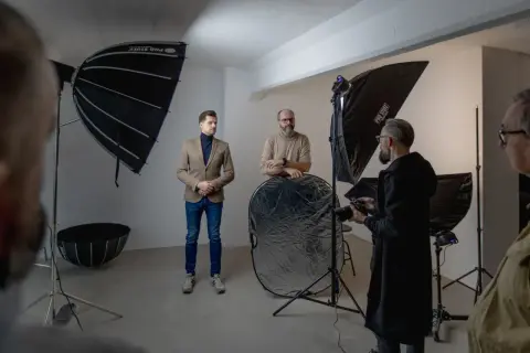 W studiu fotograficznym trwa sesja. Widać ludzi, sprzęt i modyfikatory światła w fotografii, w tym parasole i blendę.