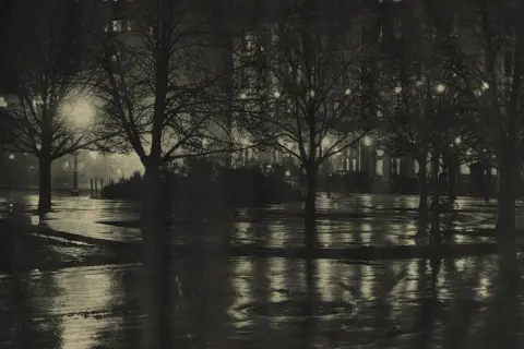 Nocny park po deszczu, drzewa bez liści, mokre ścieżki odbijające światła budynków. Alfred Stieglitz uchwycił atmosferę miasta.
