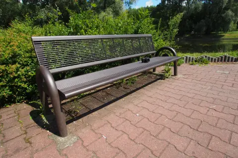 Szeroki kadr ukazuje pustą, metalową ławkę w parku, obok której leży aparat fotograficzny. W tle zielone drzewa i staw.