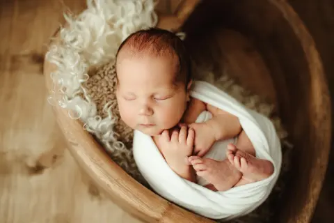 Urocze zdjęcia noworodków: śpiące maleństwo otulone białym kocykiem w drewnianej misie, otoczone miękkim futerkiem.