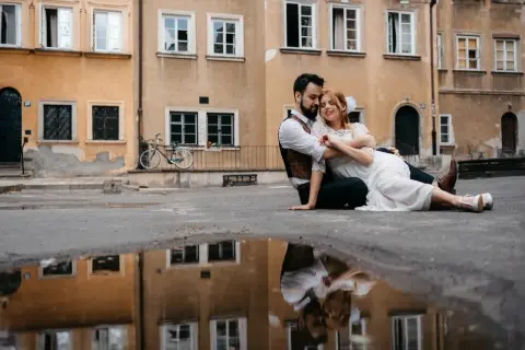 Kamil Włodarczyk i jego ukochana siedzą na ziemi, otoczeni starą kamienicą. Ich odbicie w kałuży dodaje magii tej chwili.