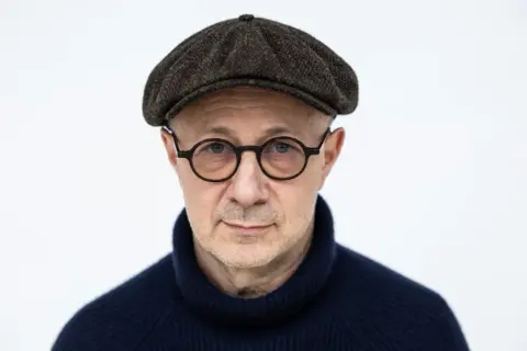 Mikołaj Grynberg w okularach i kaszkiecie, zamyślony, w granatowym golfie.
