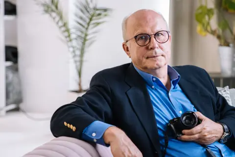Tomasz Tomaszewski, w okularach i granatowej marynarce, trzyma aparat fotograficzny.