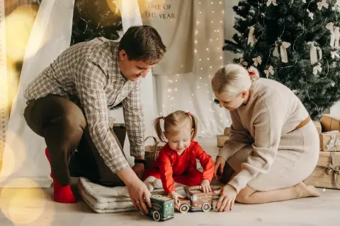 Wesoła sesja rodzinna w domu. Rodzice bawią się z córeczką zabawkami przy choince.