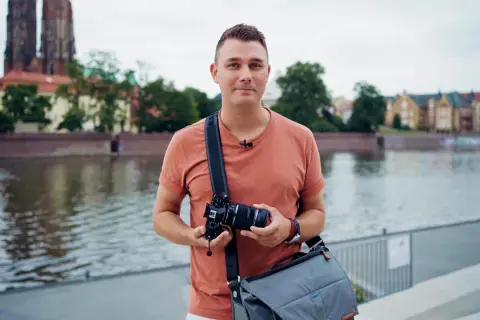 Mężczyzna z aparatem i torbą na ramieniu, gotowy do robienia zdjęć na dworze. W tle rzeka i zabytkowe budynki.
