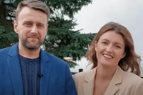 Uśmiechnięty adam kozioł w niebieskiej marynarce obok kobiety w beżowej.