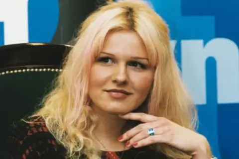 Agnieszka Lorek z blond włosami i delikatnym uśmiechem, oparta o fotel, z ręką podtrzymującą podbródek.