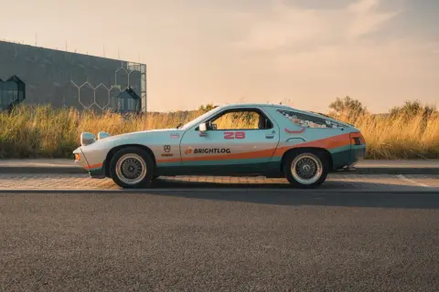 Klasyczna fotografia samochodowa: Porsche 928 w kolorach retro, z numerem 28 i logiem Brightlog, na tle nowoczesnego budynku i złotej trawy.