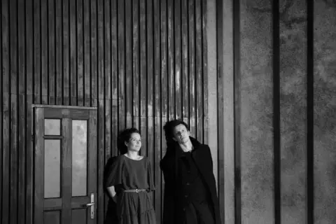 Dwoje aktorów w czerni, w dramatycznej pozie, na tle drewnianej ściany i drzwi. Ta fotografia teatralna uchwyciła moment napięcia.