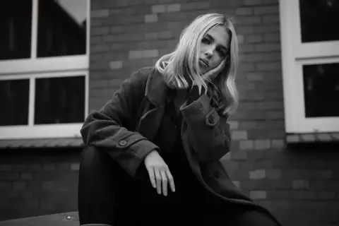 Blondynka w płaszczu opiera głowę na dłoni, zamyślona. Ten detal co to znaczy?